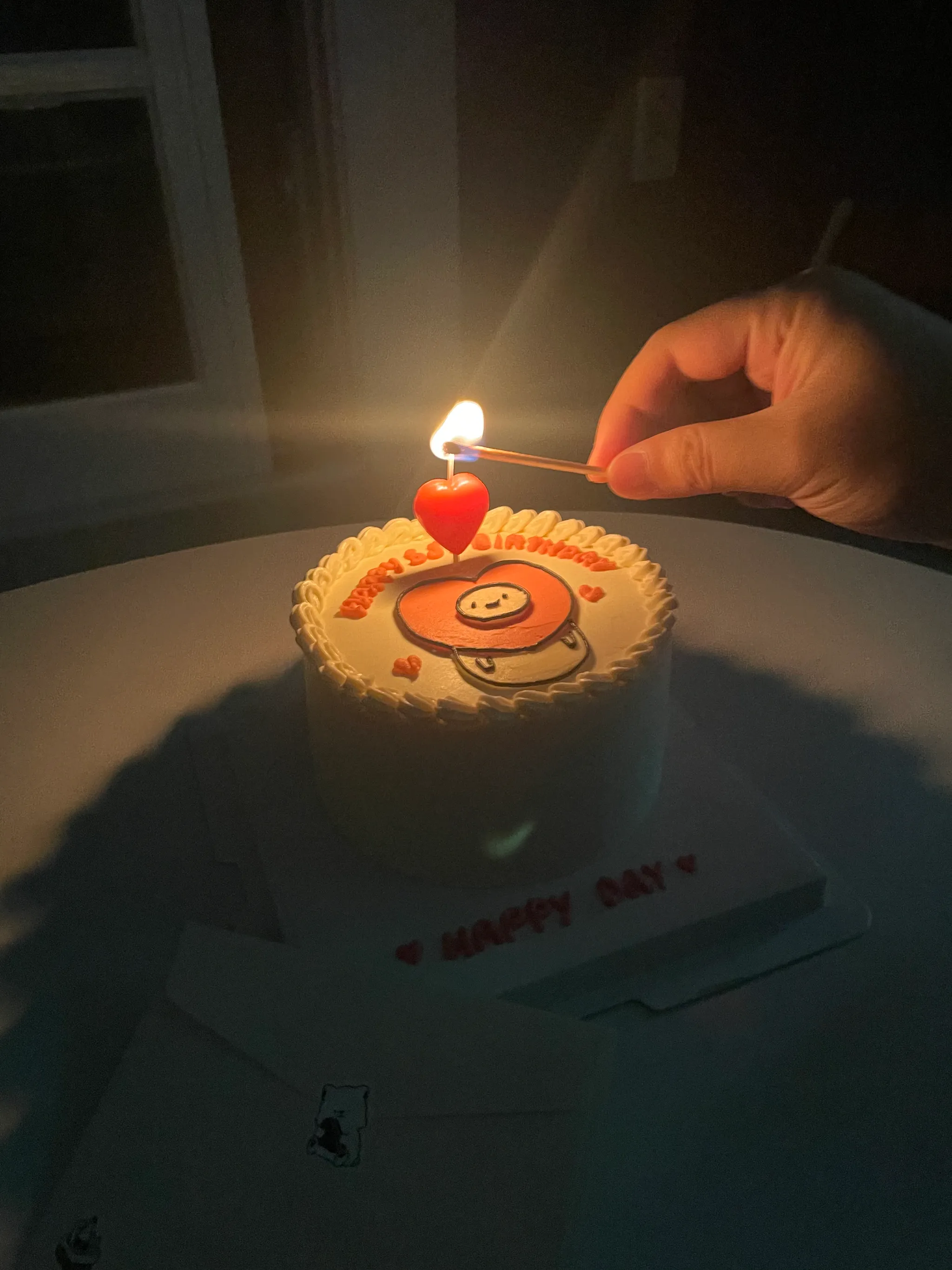 어두운 방에서 생일 케이크의 하트 모양 초에 불을 붙이는 모습.
